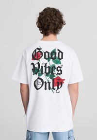 Biała koszulka z krótkim rękawem z czarnym napisem „Tylko dobre wibracje” oraz nadrukowanymi czerwonymi różami i zielonymi liśćmi na plecach. Luźny krój.