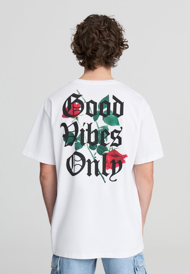 Biała koszulka z krótkim rękawem z czarnym napisem „Tylko dobre wibracje” oraz nadrukowanymi czerwonymi różami i zielonymi liśćmi na plecach. Luźny krój.
