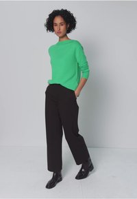 Groen gebreide trui met een geronde halslijn, gecombineerd met hooggetailleerde zwarte broek en zwarte enkellaarsjes, tegen een neutrale achtergrond.