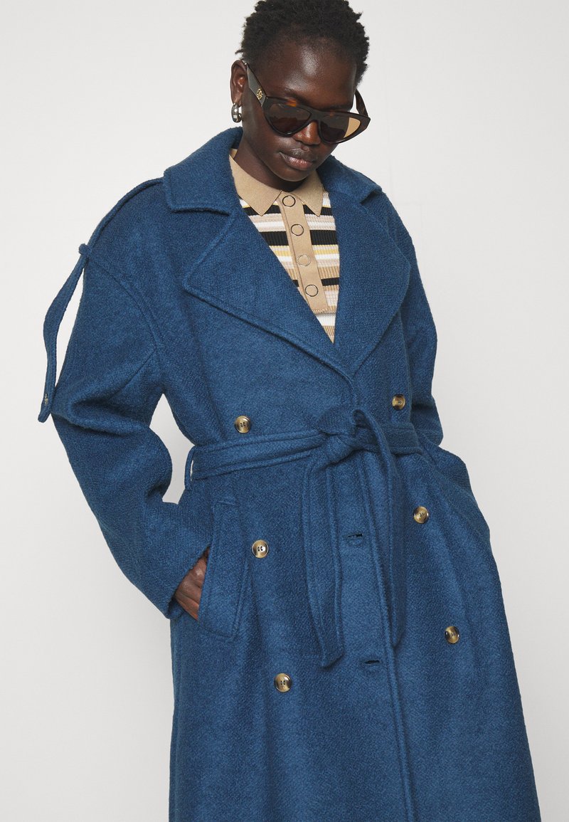 Niebieski wełniany płaszcz w mieszaninie z włóknami syntetycznymi, z paskiem w talii, podwójnym zapięciem na złote guziki, klapowym kołnierzem oraz bocznymi kieszeniami. Teksturowana tkanina.