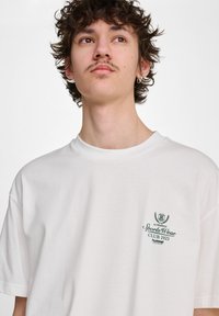 Biała bawełniana koszulka z krótkim rękawem, okrągłym dekoltem i zielonym haftowanym logo na lewej piersi z napisem "SportsWear CLUB 1923."