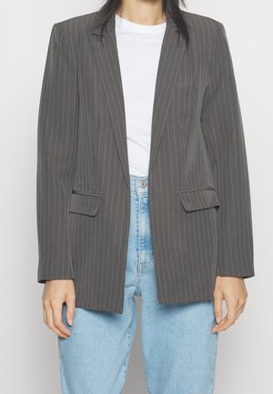 Person trägt eine dunkelgraue Nadelstreifenjacke über einem weißen Hemd und hellblauen Jeans, steht vor einem schlichten Hintergrund.