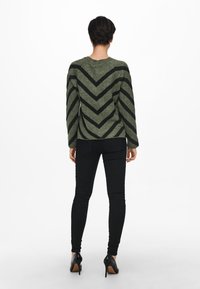 Grüner Pullover mit einem schwarzen Zickzackmuster, langen Ärmeln und rundem Ausschnitt, kombiniert mit schwarzen schmalen Jeans und High Heels.