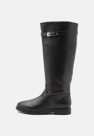 Botte haute en cuir noir avec talon bas, bride décorative et boucle argentée près du haut sur un fond blanc uni.