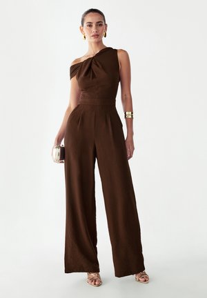 Brun, asymmetrisk jumpsuit med en skulder, der har en tætsiddende overdel, vide bukseben og en knudedetalje ved skulderen. Inkluderer et metallisk armbånd og en clutch.