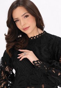 Top in pizzo nero con intricati motivi floreali, scollatura alta e maniche a campana. In risalto il dettaglio di una mano che indossa un anello decorativo.