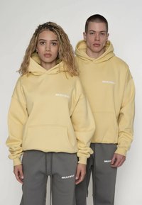 Gele hoodies met voorkantenzakken en witte "MULTIPLY" tekst, gecombineerd met grijze joggingbroeken met dezelfde merknaam. Soepele textuur.