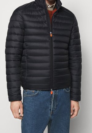 Veste matelassée noire avec un col montant, un design matelassé horizontal et une fermeture éclair avec un tirant orange. Portée par-dessus un pull et un jean.