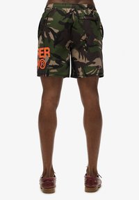 Shorts de bain camouflage avec un motif vert, marron et noir, arborant un logo orange. Fabriqués en tissu léger, ils comprennent des poches latérales.
