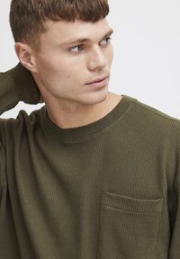 Oliven grønn langermet T-skjorte laget av teksturert stoff, med en rund hals og en brystlomme med firkantet design.