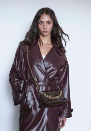 Trench coat in ecopelle bordeaux con cintura regolabile, abbinato a una clutch marrone scuro con tracolla dorata. Texture liscia.