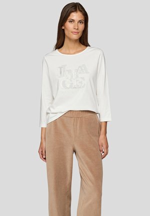 Frau trägt weißes Langarmshirt mit der Aufschrift "IMAGE" und beige Cord-Hosen mit weitem Bein vor einfachem grauem Hintergrund.