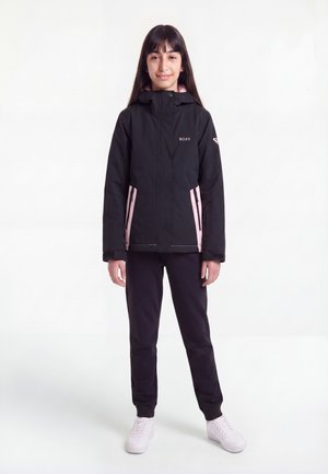 Jeune fille debout, portant une veste noire et rose zippée, un pantalon noir et des baskets blanches sur un fond blanc uni.