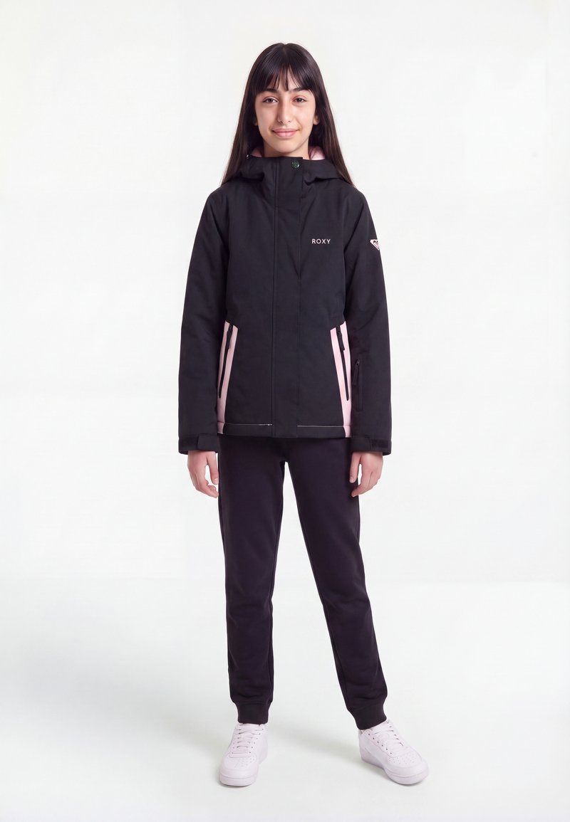 Jeune fille debout, portant une veste noire et rose zippée, un pantalon noir et des baskets blanches sur un fond blanc uni.