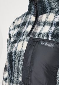 Kragefleecegenser i grå, svart og hvite mønstre; har en svart nylonlomme med Columbia-merke. Myk tekstur.