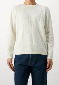 Personne portant un pull en maille blanc à manches longues avec un motif géométrique subtil et un jean bleu foncé, debout devant un fond uni.