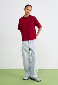 T-shirt rossa in cotone con maniche corte, abbinata a jeans in denim a gamba larga di colore azzurro chiaro. Il modello indossa sneakers grigie, in piedi su un pavimento verde.