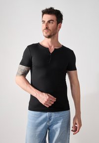 Čierne tričko s krátkym rukávom a okrúhlym výstrihom, s detailom gombíka, priliehajúce k telu, vystavené na modeli s svetlomodrými džínami.