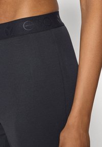 Čierne rebrované legíny s hladkým elastickým pásom, ktorý zdobí tónovaný reliéfny detail loga. Tkanina má jemnú textúru a priliehavý strih.