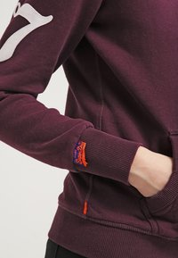 Sudadera de algodón morada con el número '7' en blanco impreso en la manga, puños acanalados y una etiqueta naranja de acento cerca del dobladillo. Textura suave.