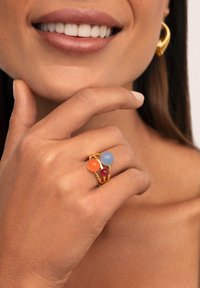 Goldener Ring mit drei gefärbten Steinen: orange, blau und rot. Der Ring hat ein verschlungenes Design mit einer glatten Oberfläche.