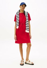 Vestito di cotone rosso con maniche corte e vita con cordoncino. Abbinato a un berretto blu navy e a un maglione a righe bianche e blu navy appoggiato sulle spalle.