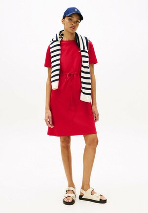 Vestito di cotone rosso con maniche corte e vita con cordoncino. Abbinato a un berretto blu navy e a un maglione a righe bianche e blu navy appoggiato sulle spalle.
