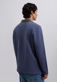 Blue long-sleeved shirt with a collared neck, featuring a contrasting gray collar and a smooth texture. Worn with denim jeans.