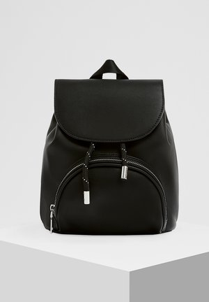 Sac à dos noir avec une poche zippée à l'avant, fermeture à cordon, rabat et poignée de transport supérieure sur une plateforme blanche.