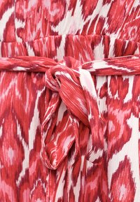 Κόκκινο και λευκό υφασμάτινο με σχέδιο, δεμένο κόμπο γύρω από τη συλλεγμένη μέση ενός ενδύματος, που δείχνει υφή και αφηρημένο σχέδιο.