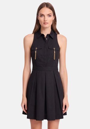 Donna che indossa un vestito nero senza maniche con colletto, tasche frontali e dettagli con nappine dorate, in piedi davanti a uno sfondo chiaro e uniforme.