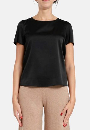 Vrouw draagt een zwarte satijnen blouse met korte mouwen en beige broek, staand met ontspannen handen langs haar zij tegen een effen achtergrond.