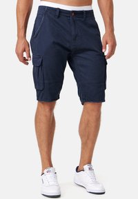 Shorts cargo bleu marine avec une coupe décontractée, dotés de poches latérales, d'une fermeture à bouton et d'emmanchures ourlées. Portés avec des baskets blanches.