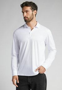 Vit långärmad polo med mjuk textur, treknappsstängning och spetsig krage, tillsammans med mörka jeans.