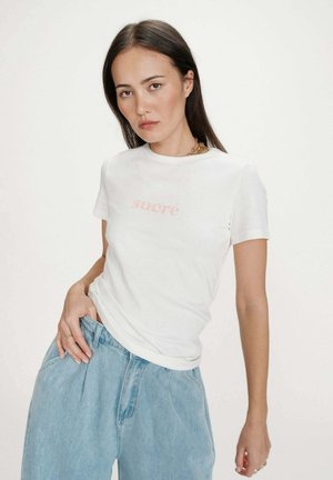 Femme aux longs cheveux noirs portant un t-shirt blanc "sucré" et un jean taille haute bleu clair, posant avec une main dans la poche devant un fond uni.
