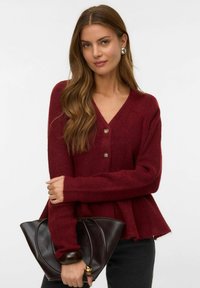 Burgundska pletena jakna s V-izrezom, gumbnim zapiranjem in peplum robom. Model drži temno rjavo torbico, kar prikazuje sproščen kroj.