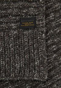 Strikket grå sweater med et struktureret mønster og lyse pletter, ribbet kant og et sort mærke med guldtekst, der siger "lavet til daglige eventyr."