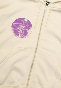 Felpa con zip beige caratterizzata da una grafica circolare viola di uno scheletro. Include due tasche frontali e un tessuto testurizzato.