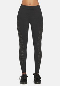 Kobieta w czarnych legginsach z ciemnymi panelami w kamuflażu po bokach i czarnych sznurowanych butach sportowych, stojąca na jednolitym tle.