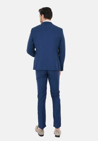Completo blu con una vestibilità su misura. La giacca ha uno spacco singolo e i pantaloni sono a taglio slim. Le scarpe sono mocassini in suede grigio chiaro.