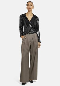 Schwarze Paillettenbluse mit Kragen, kombiniert mit braunen Hochwasser-Hosen mit weitem Bein. Schwarze spitz zulaufende Absätze runden das Outfit ab.