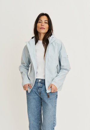 Vrouw die een lichtblauw capuchonjack, een wit shirt en blauwe spijkerbroek draagt, staand tegen een effen lichte achtergrond.