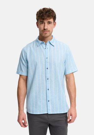 Mężczyzna w krótkim rękawie, jasnoniebieskiej koszuli zapinanej na guziki z pionowymi białymi paskami i ciemnych spodniach na jednolitym tle.