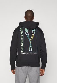Sweatshirt preto com um gráfico de mosquetões de escalada nas costas. Feito de tecido macio, possui um bolso canguru e capuz com cordão.