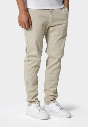 Hombre con pantalones beige ajustados y zapatillas blancas, de pie con las manos relajadas, visible desde la cintura hasta los pies sobre un fondo liso.