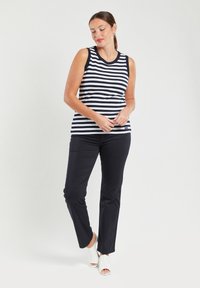 Pasek bezrękawnik w granatowo-białym materiale, zestawiony z czarnymi spodniami. Modelka nosi białe sandały na obcasie i prezentuje neutralne tło.