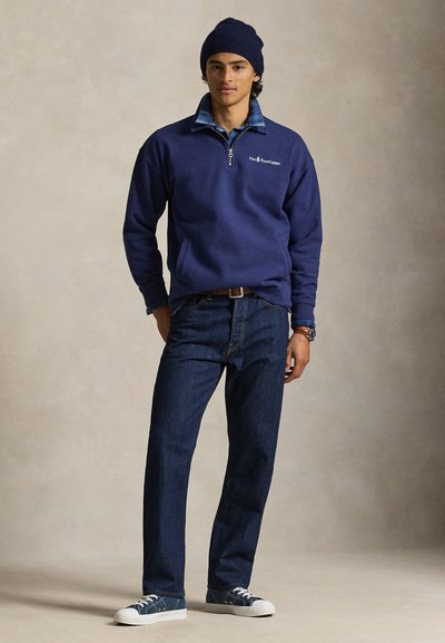 Νέος άνδρας που φοράει μπλε σκουφάκι, μπλε φούτερ με φερμουάρ μέχρι το στέρνο, από πάνω από καρό πουκάμισο, σκούρα τζιν και μπλε αθλητικά παπούτσια, στέκεται μπροστά σε απλό φόντο.