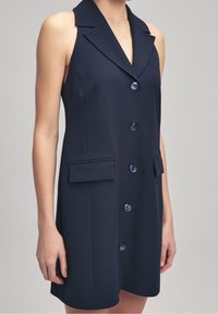 Robe bleu marine sans manches avec col à revers, boutons sur le devant et deux poches à rabat portée par une personne sur un fond uni.