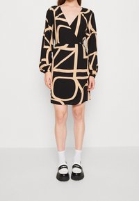 Schwarzes Kleid mit tiefem V-Ausschnitt, langen Ballonärmeln und beigen abstrakten Mustern. Kombiniert mit weißen Socken und schwarzen Plateau-Schuhen.