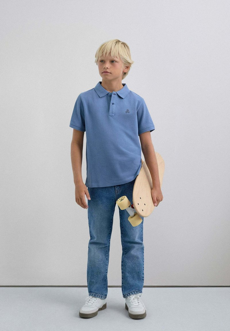 Lyseblå polo skjorte med krave, parret med blå denimbukser. Holder en træskateboard med sorte hjul. Hvide sneakers.